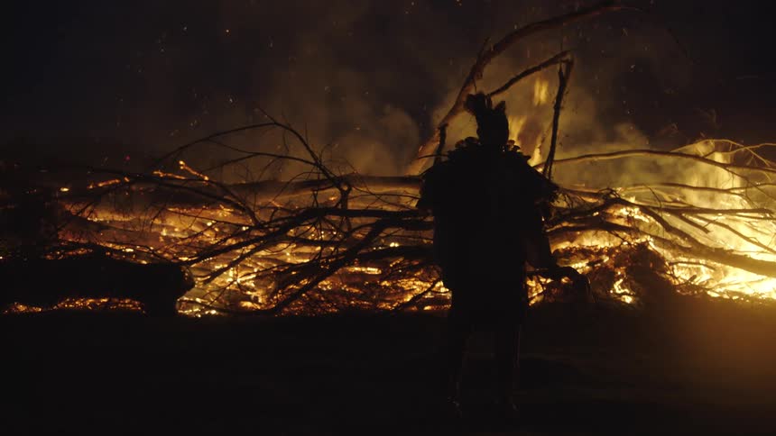 An indigenous man in a feathered headdress stands as a dark silhouette against a massive bonfire of tangled branches that burns intensely under the night sky while smoke and sparks rise into the air