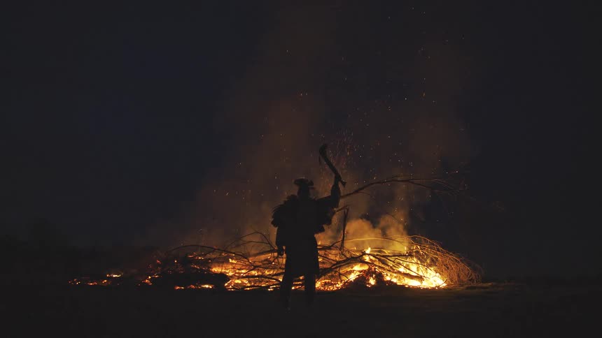 A silhouetted figure in traditional attire stands before a large crackling bonfire at night raising an axe toward the sky sparks and smoke drift into the dark air as the flames illuminate the figures dramatic pose against the wilderness
