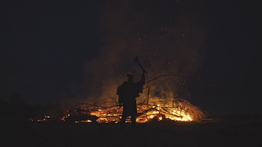A man stands in silhouette before a large bonfire at night and raises a tool into the air as orange sparks and thick smoke rise into the dark sky
