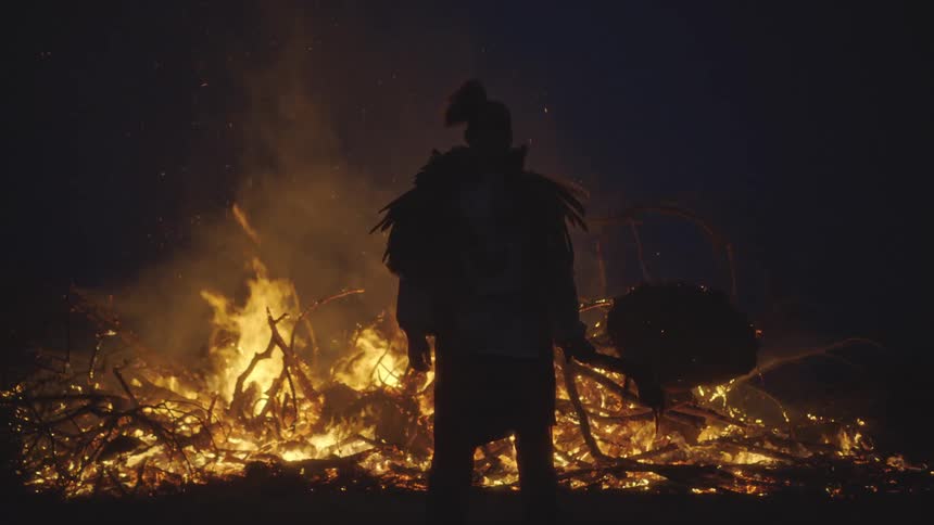 Figure in ceremonial attire holding drum before bonfire