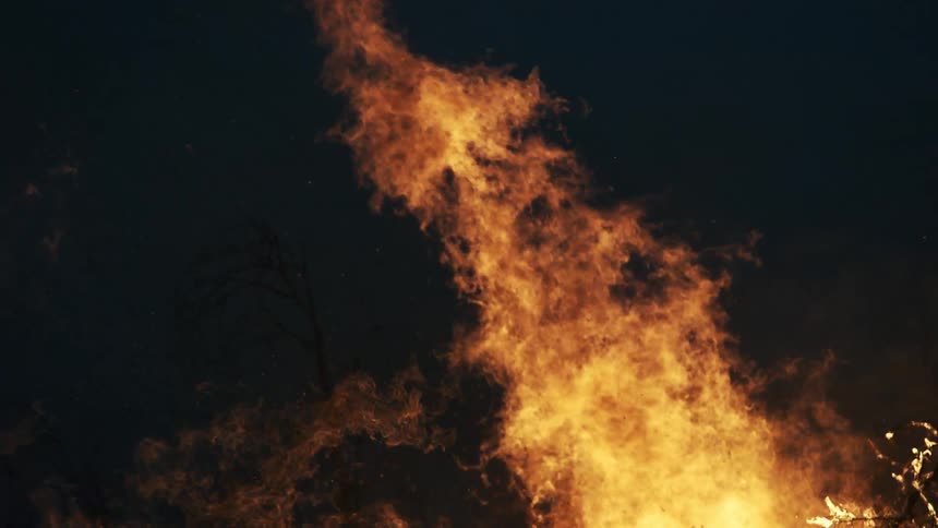 A close view of bright orange flames and thick gray smoke rising from a large bonfire against a dark night sky with small sparks drifting through the air