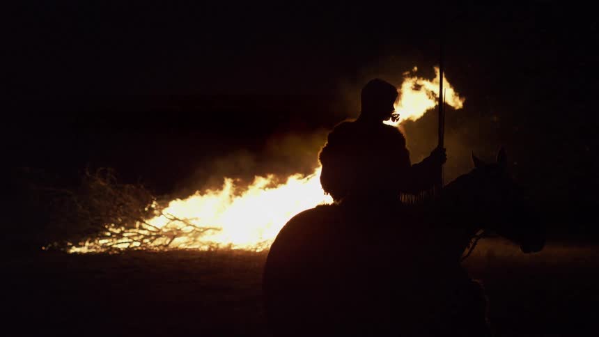 A man on a horse sits in silhouette before a large bonfire at night holding a long staff as bright orange flames rise from a pile of branches into the dark sky