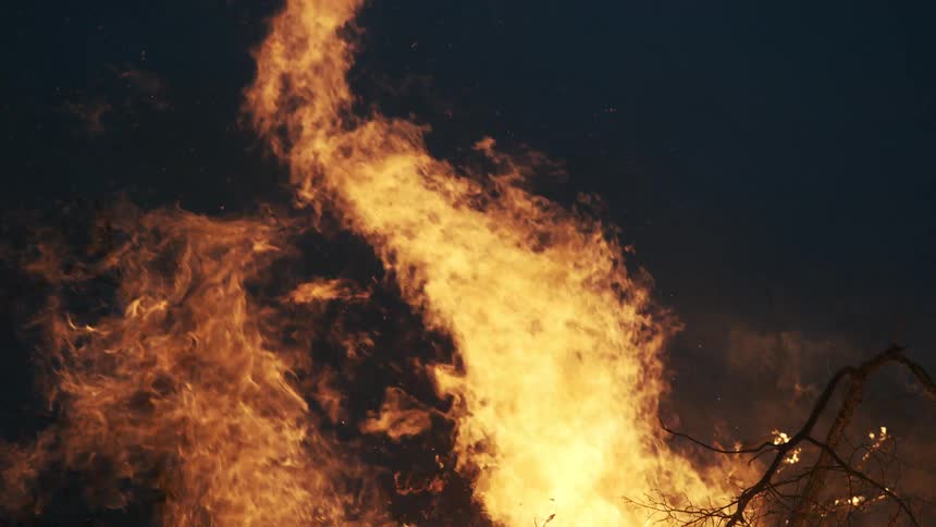 A close view of a large bonfire with bright orange flames consuming dry branches and sending small sparks into the dark night sky