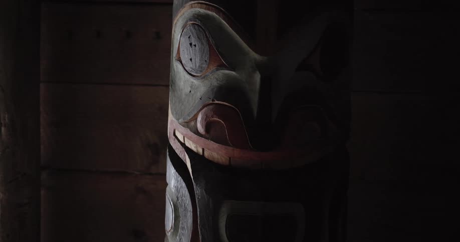 A closeup of a handcarved totem pole inside a dimly lit wooden structure light from an unseen source on the left side slowly sweeps across the intricate carvings revealing the weathered textures and traditional pigments