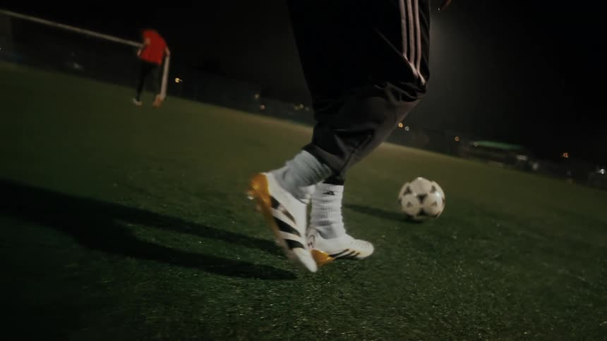 A close view shows the feet of a person wearing white and gold soccer cleats as they dribble a soccer ball across a green turf field at night