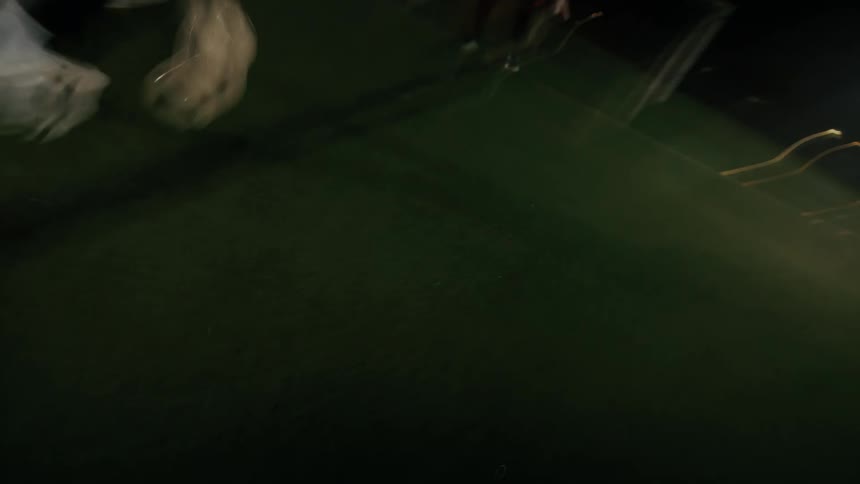 A soccer player in white cleats and black pants dribbles a ball across a green turf field at night toward a goal post while the camera follows the fast movement from a low angle