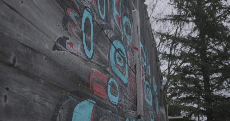A closeup of a wooden wall features bold blue and red painted designs with flurries falling on tall evergreen trees visible in the background