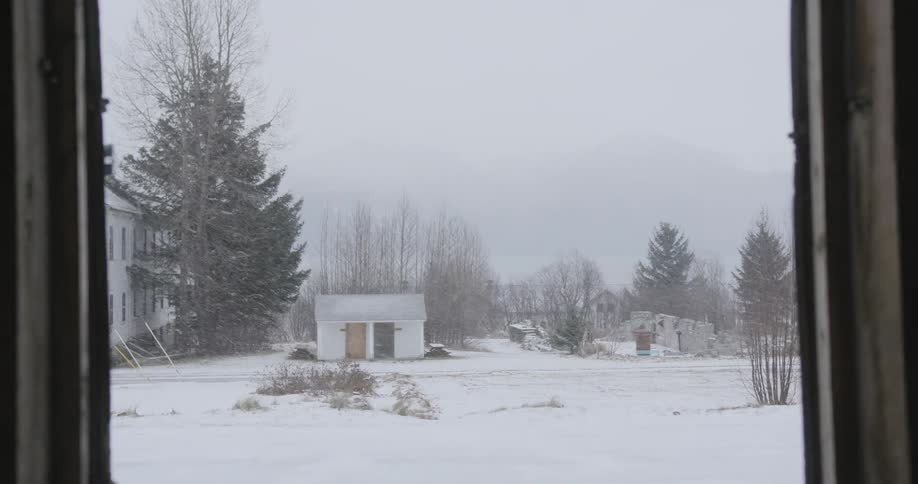 A view from inside a dark building through opening wooden doors shows a small white shed in a snowy field with trees and a larger building during a heavy winter snow storm