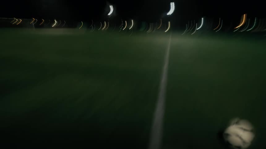 A motionblurred figure in a red shirt runs across a lit field at night with arms raised trying to control a soccer ball