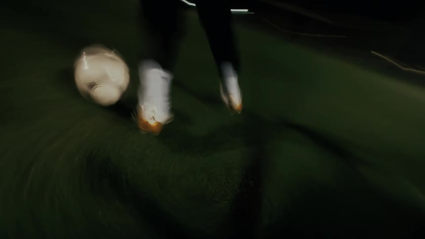 A person in white socks and cleats dribbles a soccer ball across an artificial turf field at night under bright stadium lights