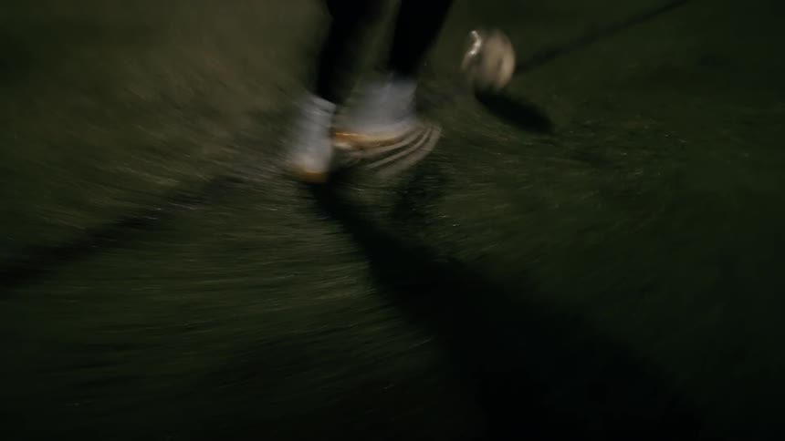 A soccer player in white cleats and black pants dribbles a ball across an artificial turf field at night under bright stadium lights while the camera follows the fast footwork from a low angle