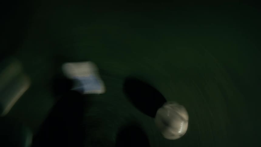 An overhead view shows the feet of a person in white sneakers and dark pants as they dribble a white soccer ball across a green turf field at night