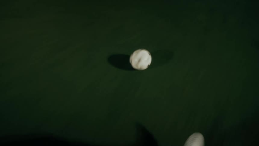 An overhead view shows the feet of a person in white sneakers and dark pants as they dribble a white soccer ball across a green turf field at night