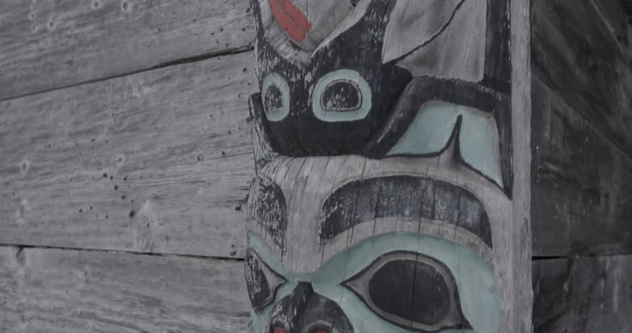A close view shows the intricate carved faces and figures of a wooden totem pole painted in red and black attached to the side of a weathered wooden building
