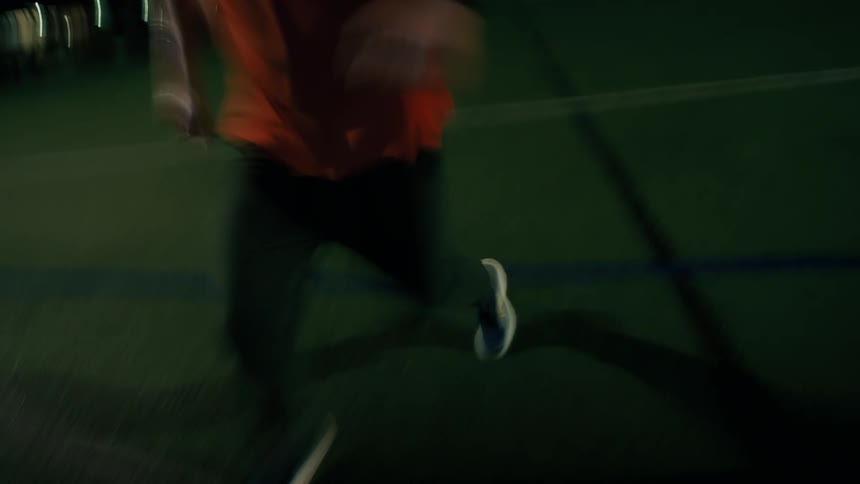 An overhead view shows the feet of a person they dribble a white soccer ball across a green turf field at night