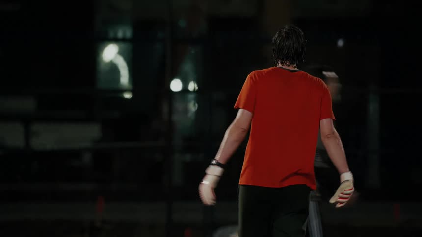 A man wearing an orange shirt and goalie gloves stands on a soccer field at night while other players move past him in the background under bright lights