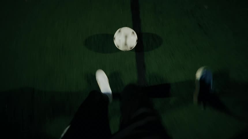An overhead view shows the feet of a person in white sneakers and dark pants as they dribble a white soccer ball across a green turf field at night