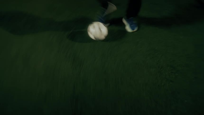 A person wears blue sneakers and dark pants while dribbling a white soccer ball across a green artificial turf field at night