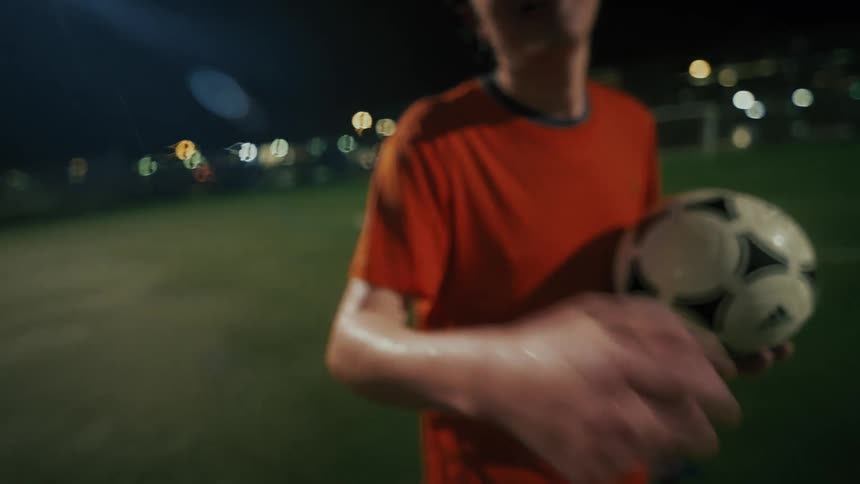 A man wearing an orange shirt stands on a green sports field at night holding a soccer ball and gives a fist bump to the camera