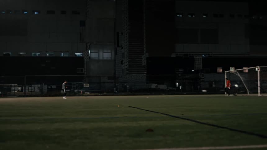 Two players on a field at night one standing as the goalkeeper while the other kicks the ball toward him and he blocks it with his feet