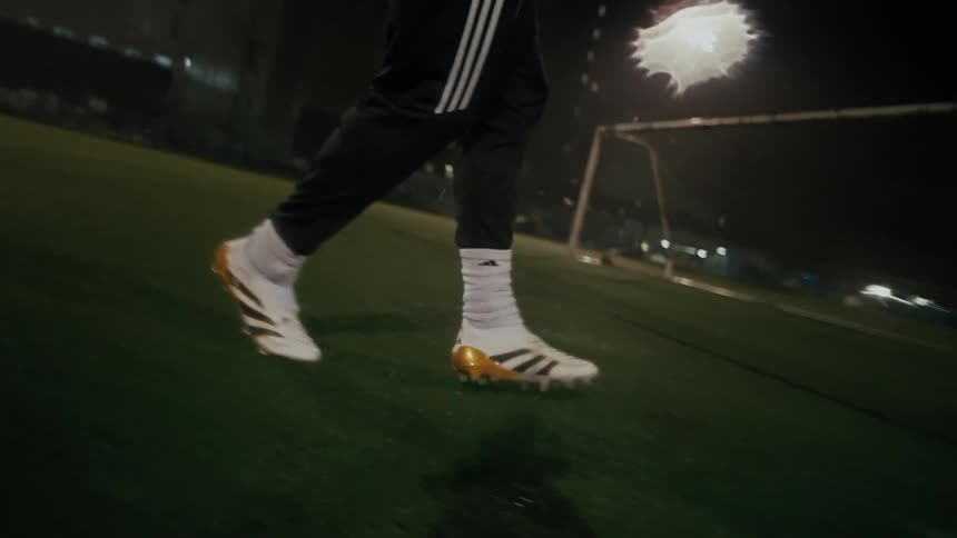 A soccer player wearing white and gold cleats practices footwork and ball control on a dark rainy field at night the closeup footage captures the athletes movements across the turf with a goal post and stadium lights visible in the background