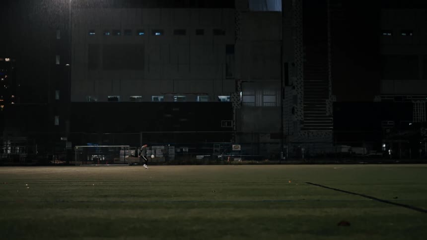 A distant view shows a soccer player kicking a ball across a green field at night toward a goal where a goalie stands ready to block the shot