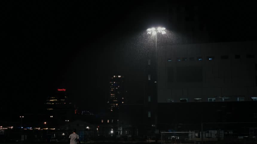 Under bright stadium lights in the rain a lone man runs on a soccer field at night with city buildings glowing in the background