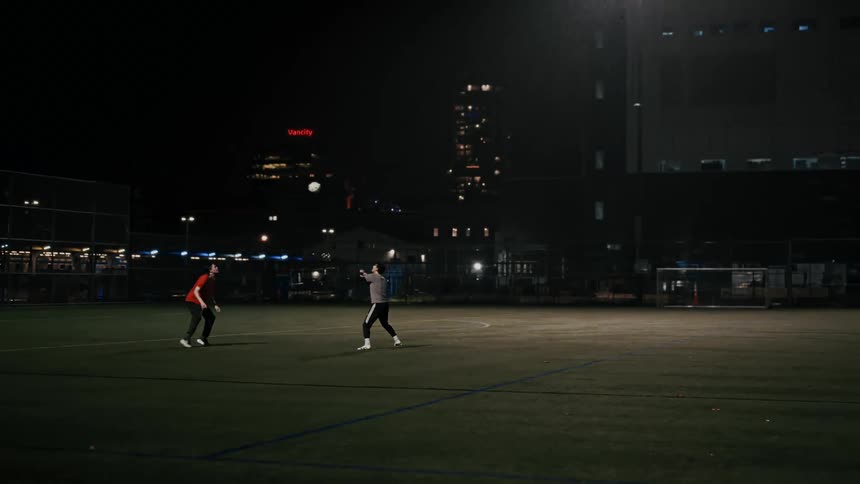 Two friends practice soccer on a green turf field at night as one man tosses the ball into the air while the other man prepares to head it back in front of a dark city skyline