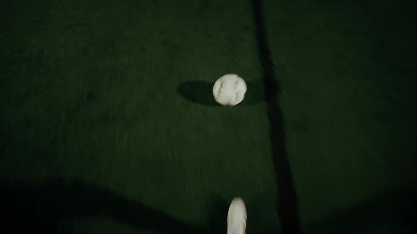 An overhead closeup shows a player in white cleats controlling a soccer ball on a dark green field under bright night lights