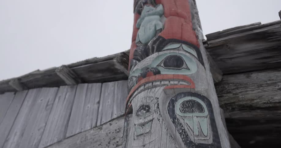 A wooden totem pole with carved faces and figures painted in red teal and black stands against a weathered wooden building as snow falls lightly under an overcast sky