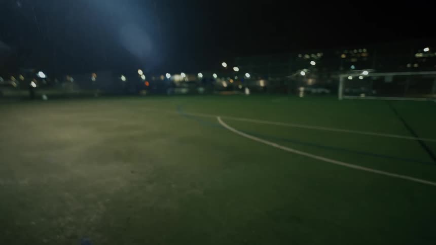 A man jogs across a brightly lit soccer field at night passing the ball to someone as they fist bump just before the camera sweeps across the empty green