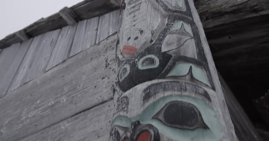 A tall totem pole carved and painted with bold red black and turquoise figures is shown from top to bottom against a gray sky extending down to the wooden structure at ground level
