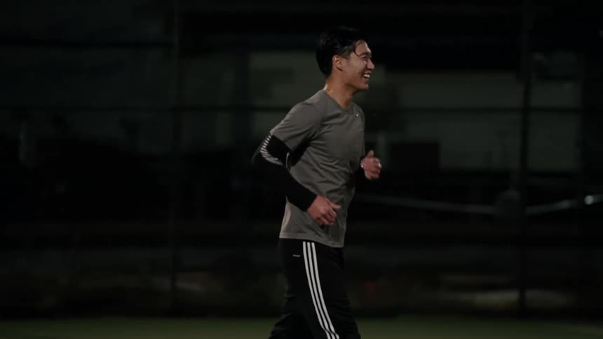 A man wearing a grey athletic shirt and black pants with white stripes jogs across a green sports field while smiling during the night