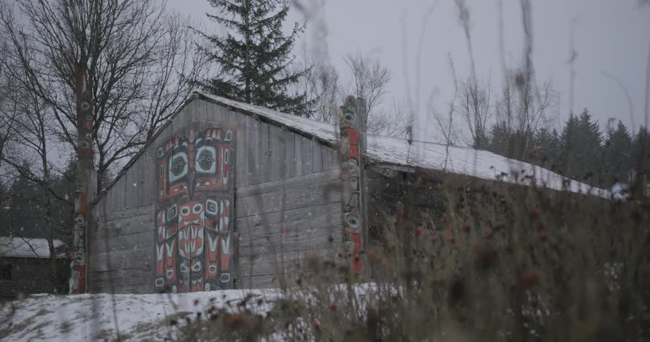 The video shows a traditional indigenous longhouse with large colorful formline artwork painted on its front doors during a light snowfall two carved totem poles stand on either side of the building which is surrounded by a winter landscape of bare tre