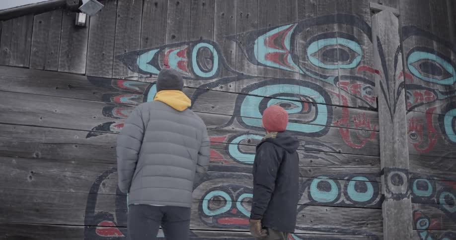 Two people stand outside during winter as they look at a large indigenous mural painted on the side of a weathered wooden building