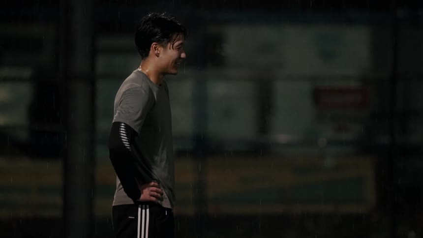 A smiling young man in athletic wear stands in the pouring rain looking off to the side with his hand on his hipthe scene is set at night on a sports court with the rain illuminated against the dark blurred background