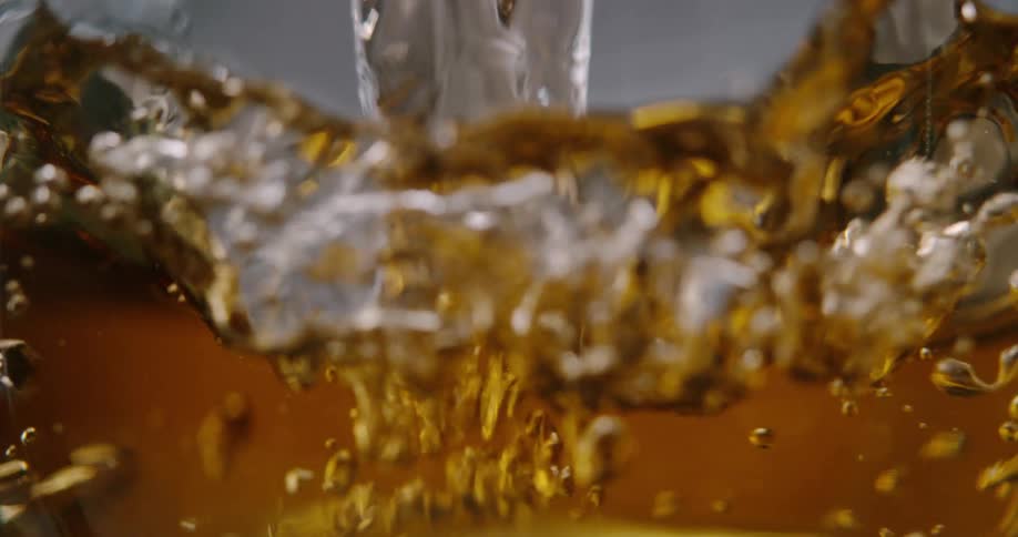 A stream of golden amber liquid pours into a glass container creating a violent splash and a swirl of bubbles
