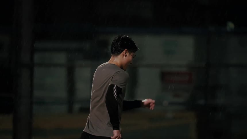 A soccer player jogs across a dark rainy field before striking a ball with his right foot the camera follows the action from a side profile focusing on his movement and the impact of the kick