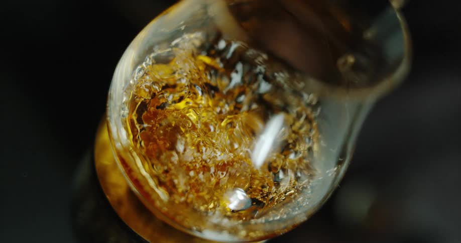 An amber liquid pours into a glass snifter from a high angle causing a golden splash