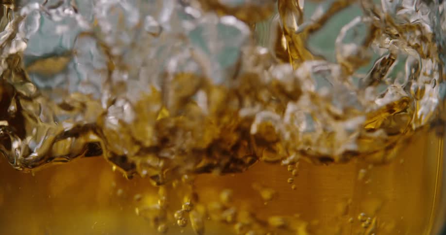 An amber liquid likely whiskey or apple juice is vigorously poured into a glass creating a splash of golden wavesthe movement produces a swarm of tiny bubbles and turbulent ripples that fill the frame in a closeup slowmotion shot
