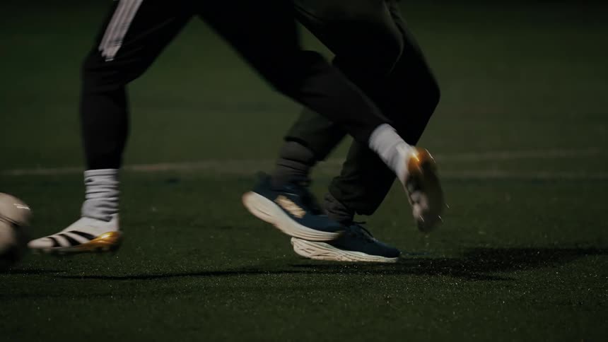 A closeup of players feet as they move a soccer ball across a dark turf field one player dribbles the ball before it is intercepted or passed with both individuals wearing athletic pants and sneakers