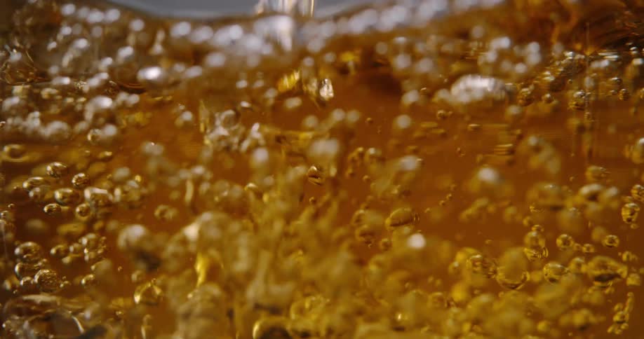 Golden liquid is poured into a glass creating a turbulent rush of bubbles and splashes the slowmotion closeup captures the amber fluid churning as it fills the frame with effervescence