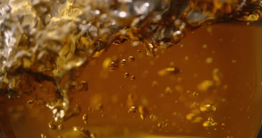 An object falls into a glass of ambercolored liquid creating a large splash and a swirl of golden bubbles the liquid continues to churn and fill with carbonation as the surface settles