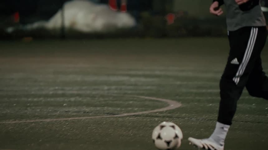 A man wearing a gray shirt and black pants runs across a green turf field at night and begins to dribble a white soccer ball with his feet in a sports facility
