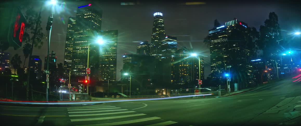A time lapse shows streaks of light from cars moving through a city intersection at night with tall illuminated skyscrapers and bright street lamps in the background
