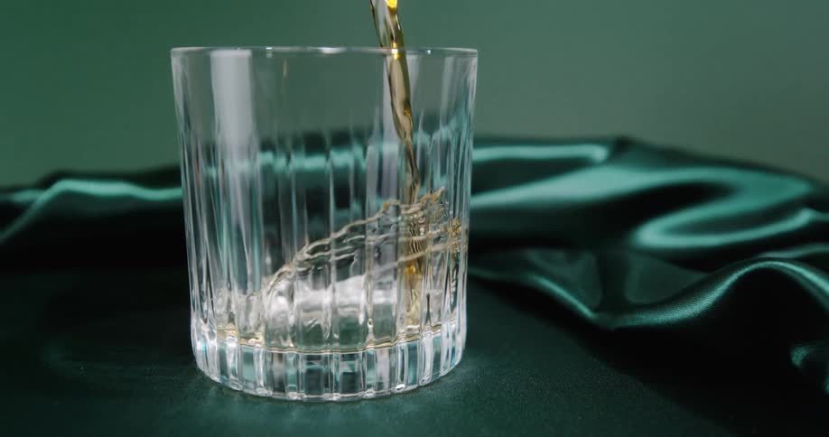 Amber whiskey pours from the neck of a dark glass bottle into a ribbed crystal glass