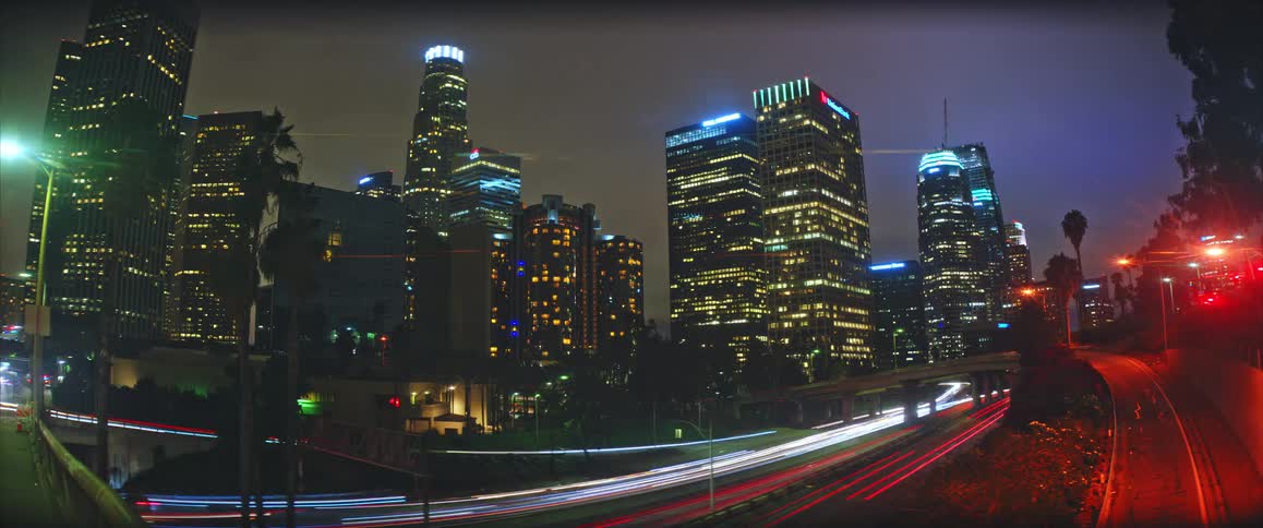 A wide view of a city skyline at night features bright lights from tall buildings while streaks of white and red light from moving traffic pass along a highway in the foreground under a dark sky