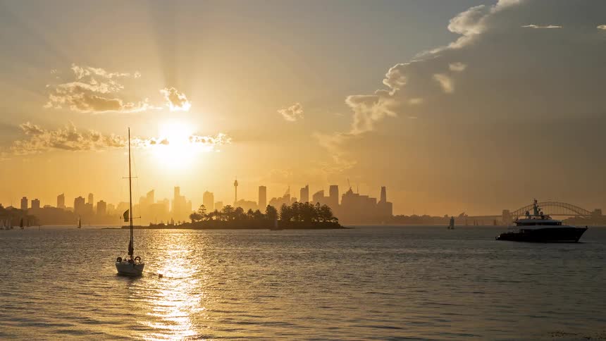 A golden hour timelapse of the sydney skyline featuring the harbor bridge and silhouetted sailboats drifting across the water as the sun dips lower the sky transitions into deep orange hues while light rays break through the clouds