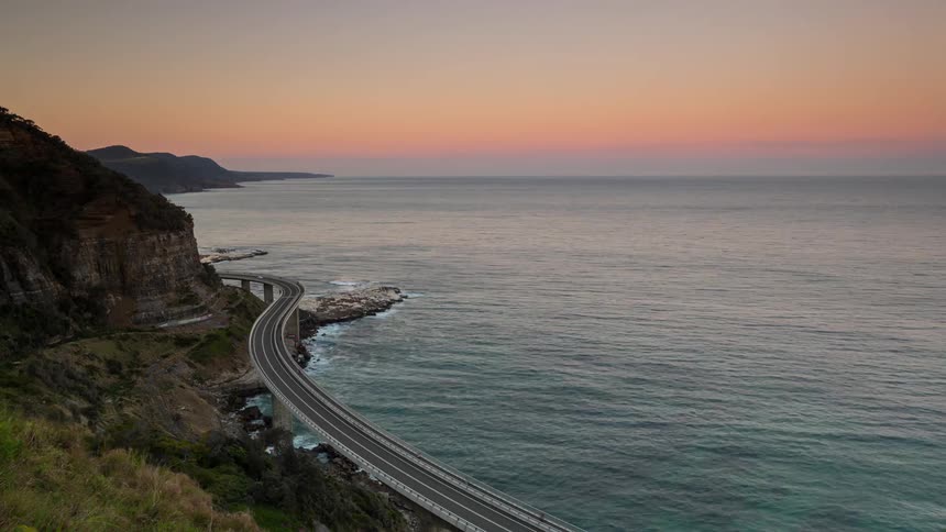 A timelapse captures the sunset fading over a winding coastal highway that curves alongside the vast open ocean