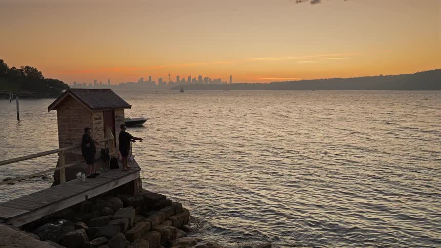 People fishing on the coast as boats sail on the water with buildings on metropolitan skyline in the distance as the sun sets